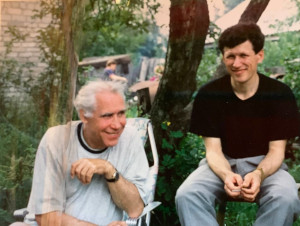 Two men, one older, one younger, sitting on chairs outdoors surrounded by grass and trees, both smiling