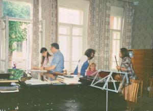 A man and four women, in a room surrounded bu musical instruments. They all appear very focused