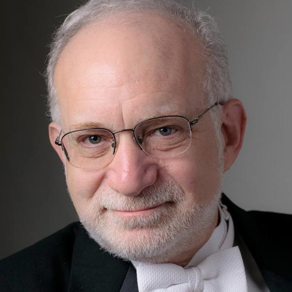 A grey-haired man wearing glasses and a black suit with a white bow tie.