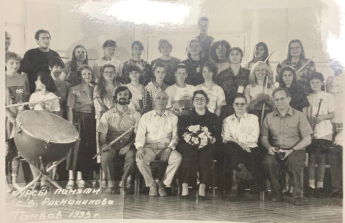 A black-and-white group photo with three rows of people, many holding flutes. A large drum sits next to the front row. Terry sits in the center of the front row, holding a bouquet of flowers