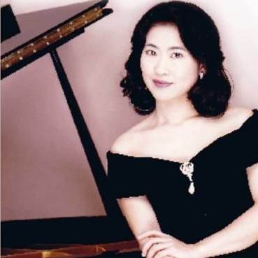 A dark-haired woman in a black dress standing gracefully beside a piano, half-smiling