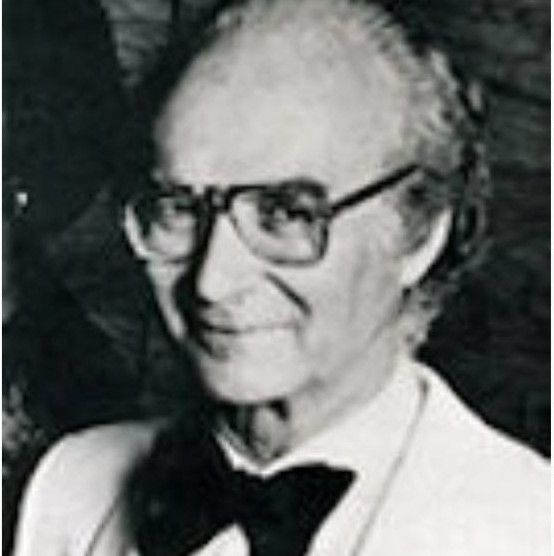 A black-and-white photo of a man with large glasses, wearing a white suit and large black bowtie. He is holding a flute.