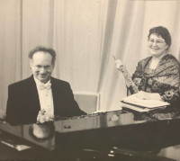 A black-and-white photo of a man and woman dressed in formal attire sitting at a piano, playing music together while the woman holds a flute