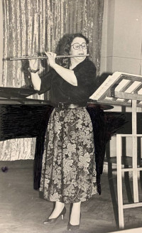 A black-and-white photo of a woman dressed formally in a black shirt and a long flowered skirt. She is standing playing the flute in front of a music stand